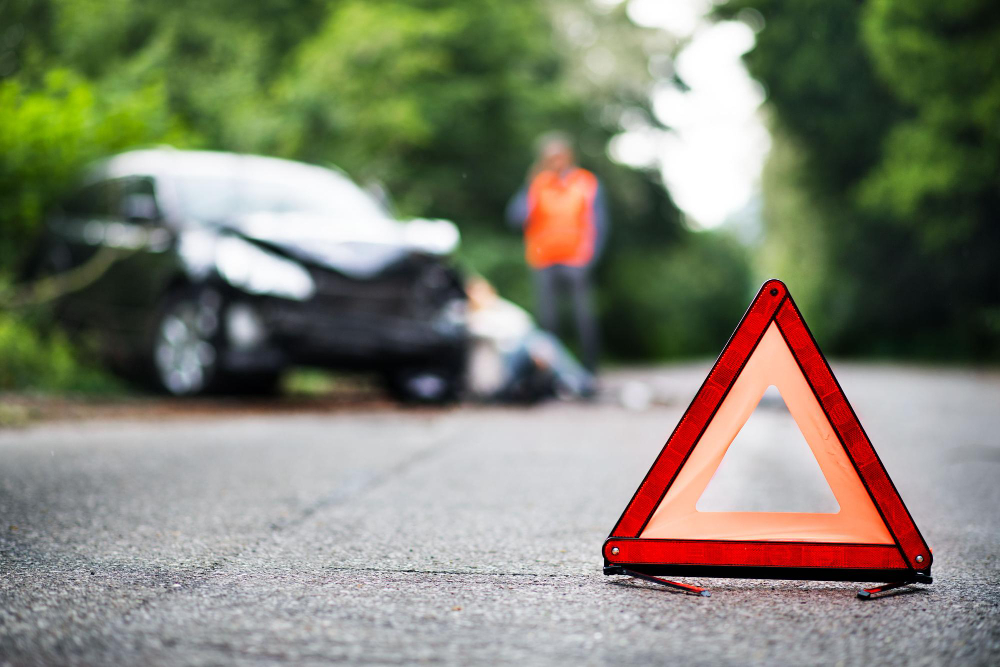 Sinal de triângulo de segurança na pista após acidente de trânsito em estrada, com carro danificado e pessoas ao fundo,