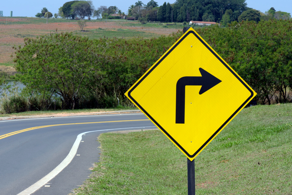 Placa de sinalização viária em fundo amarelo indicando uma curva acentuada à direita. A placa está instalada à margem de uma estrada asfaltada em área rural, com vegetação e campos ao fundo.